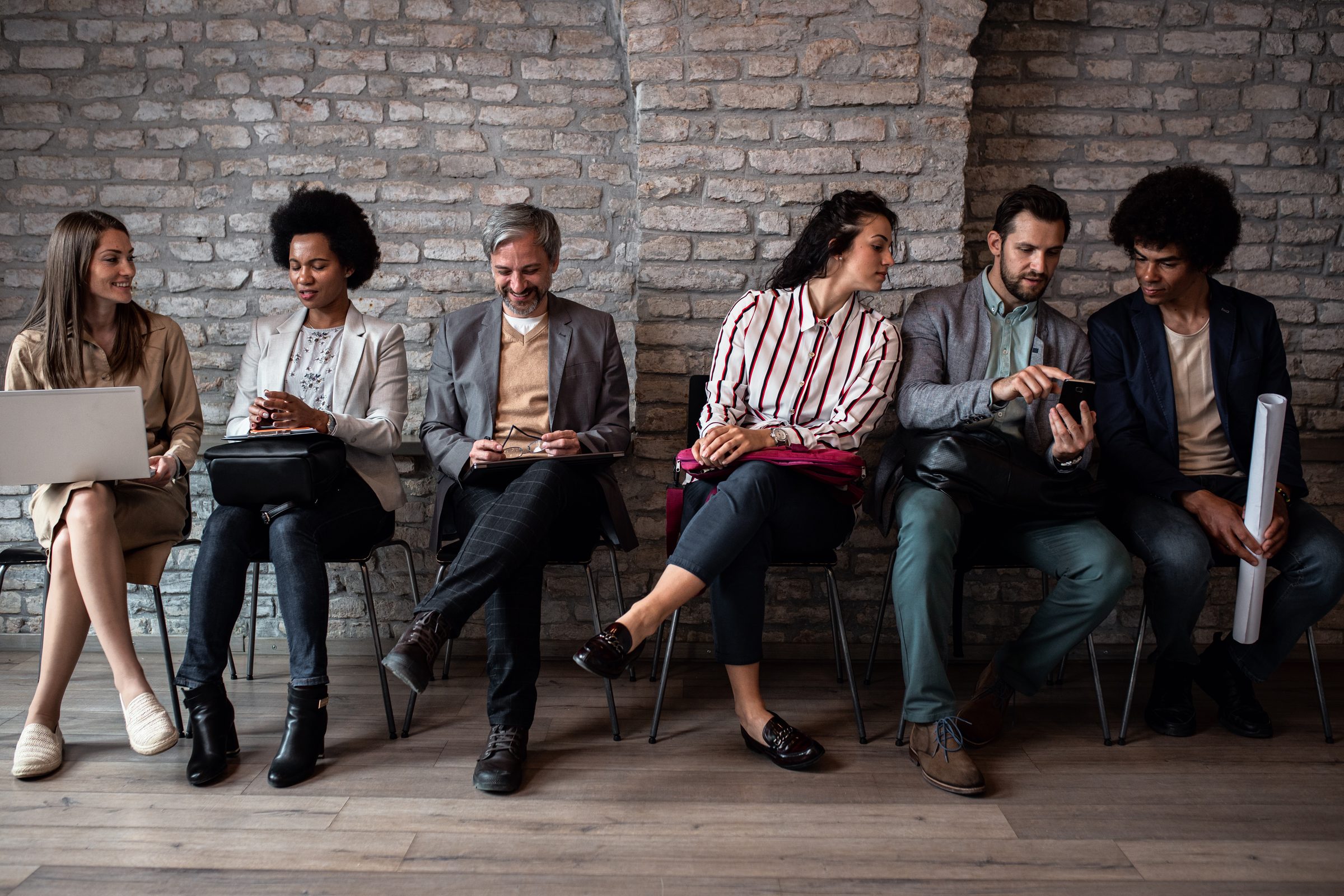 Six candidates waiting in a row of chairs against a brick wall before a casting call